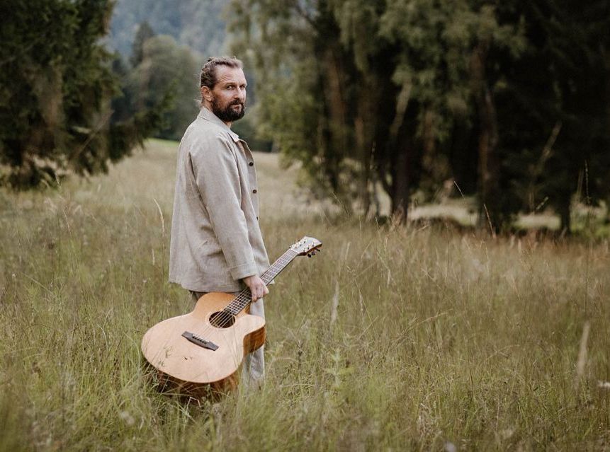Ein Mann mit Bart steht mit einer Akustikgitarre in der Hand auf einer Wiese und trägt eine helle Jacke. Im Hintergrund sind Bäume und ein Waldgebiet zu sehen.