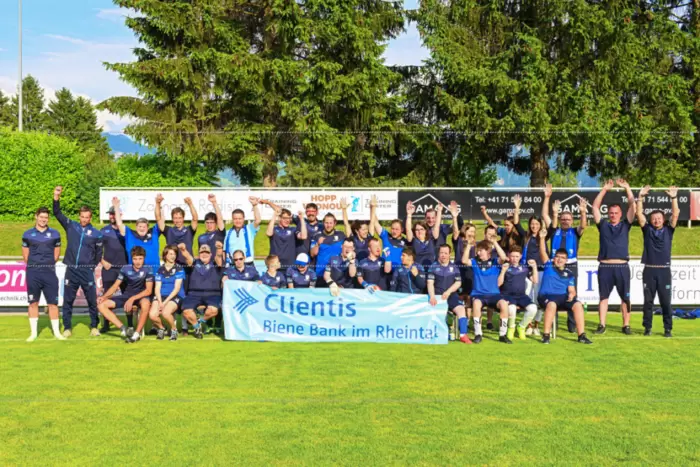Eine Fußballmannschaft in blauen Trikots feiert auf einem Rasenfeld, posiert gemeinsam und hält ein Transparent mit der Aufschrift „Clientis Biene Bank im Rheintal“ hoch, im Hintergrund sind Bäume und Werbetafeln zu sehen.