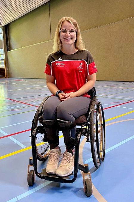 Romy Tschopp sitzt in Sportbekleidung in der Mitte einer Turnhalle und lächelt in die Kamera..