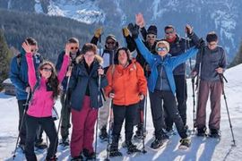 Eine Gruppe von neun Personen in Winterkleidung und Schneeschuhen posiert fröhlich auf einem verschneiten Bergpfad, hebt die Arme und lächelt mit verschneiten Gipfeln und Kiefern im Hintergrund.