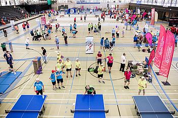 Eine große Sporthalle, in der Gruppen von Menschen Tischtennis und andere Spiele spielen. Die Teilnehmer tragen bunte Trikots, und es sind Veranstaltungsbanner zu sehen. Zuschauer und Spieler füllen den hellen, belebten Raum.