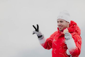 Ein lächelnder Sportler in roter Jacke und weißer Mütze hält mit einer Hand eine Goldmedaille hoch und macht mit der anderen ein Friedenszeichen vor einem schlichten, hellen Hintergrund.