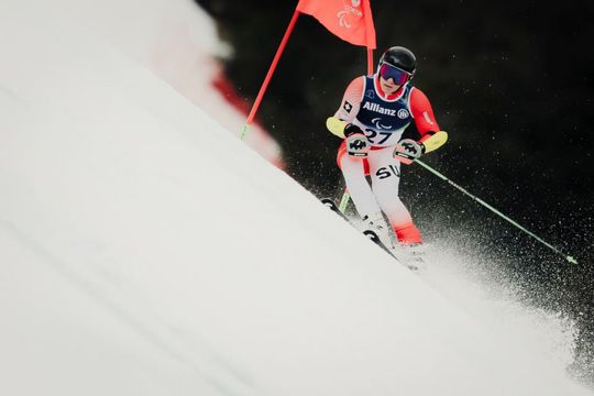 Ein Skifahrer in einem rot-weißen Anzug rast mit Helm und Brille auf einer verschneiten Piste an einer roten Fahne vorbei bergab, während der Schnee von den Skiern spritzt.