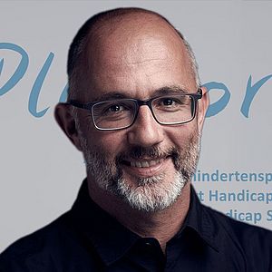 Ein Mann mit Brille und Bart lächelt in die Kamera. Er steht vor einer Kulisse mit den Aufschriften „PluSport“, „Behindertensport Schweiz“, „Sport Handicap Suisse“ und „Sport Handicap Svizzera“ sowie einem Schweizerkreuz als Logo.
