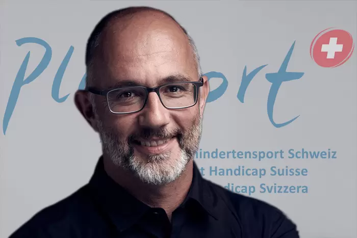 Ein Mann mit Brille und Bart lächelt in die Kamera. Er steht vor einer Kulisse mit den Aufschriften „PluSport“, „Behindertensport Schweiz“, „Sport Handicap Suisse“ und „Sport Handicap Svizzera“ sowie einem Schweizerkreuz als Logo.