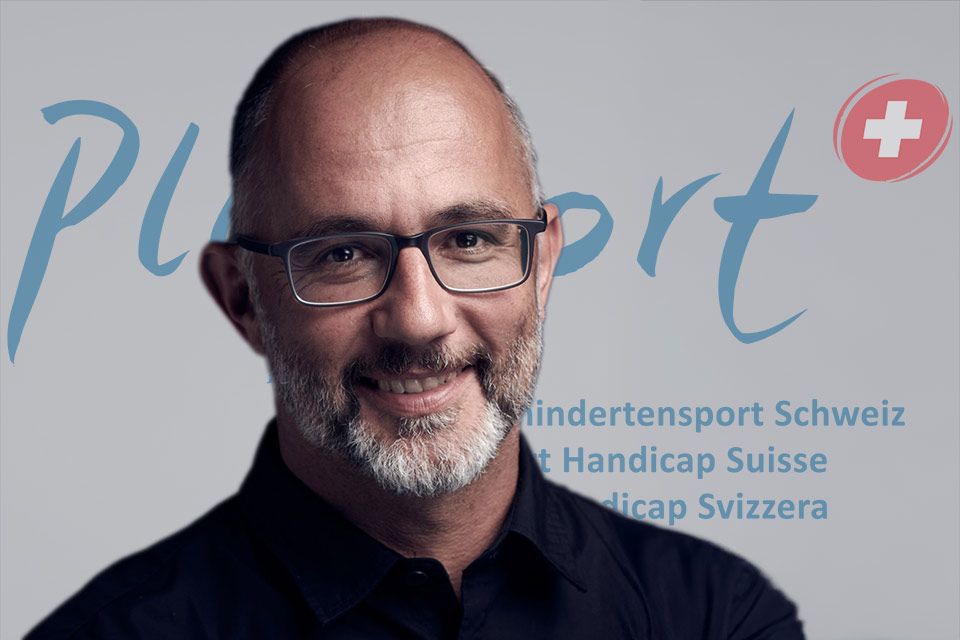 Ein Mann mit Brille und Bart lächelt in die Kamera. Er steht vor einer Kulisse mit den Aufschriften „PluSport“, „Behindertensport Schweiz“, „Sport Handicap Suisse“ und „Sport Handicap Svizzera“ sowie einem Schweizerkreuz als Logo.