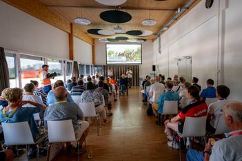 Eine Gruppe von Personen sitzt während einer Präsentation in Reihen vor einem Bildschirm im vorderen Teil eines hellen, modernen Raums. Ein Redner steht auf einem Podium, und ein Kameramann zeichnet die Veranstaltung auf. Auf dem Bildschirm wird eine Folie über das Radfahren gezeigt.