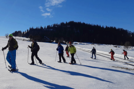 Eine Gruppe von Menschen in Winterkleidung geht mit Skistöcken und Schneeschuhen durch eine verschneite Landschaft unter einem klaren blauen Himmel, mit Bäumen und einem Gebäude im Hintergrund.