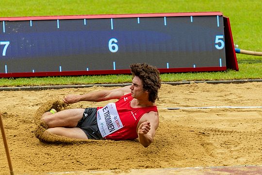 Ein männlicher Athlet in einem roten Trikot landet während eines Weitsprungwettbewerbs in einer Sandgrube. Im Hintergrund ist auf einer Rasenfläche eine Messtafel zu sehen.
