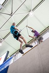 Eine Person in einem Klettergurt seilt sich an einer Indoor-Kletterwand ab, während eine andere Person von oben hilft und das Seil hält. Beide wirken konzentriert, und im Hintergrund sind Metallgeländer und ein begrüntes Dach zu sehen.