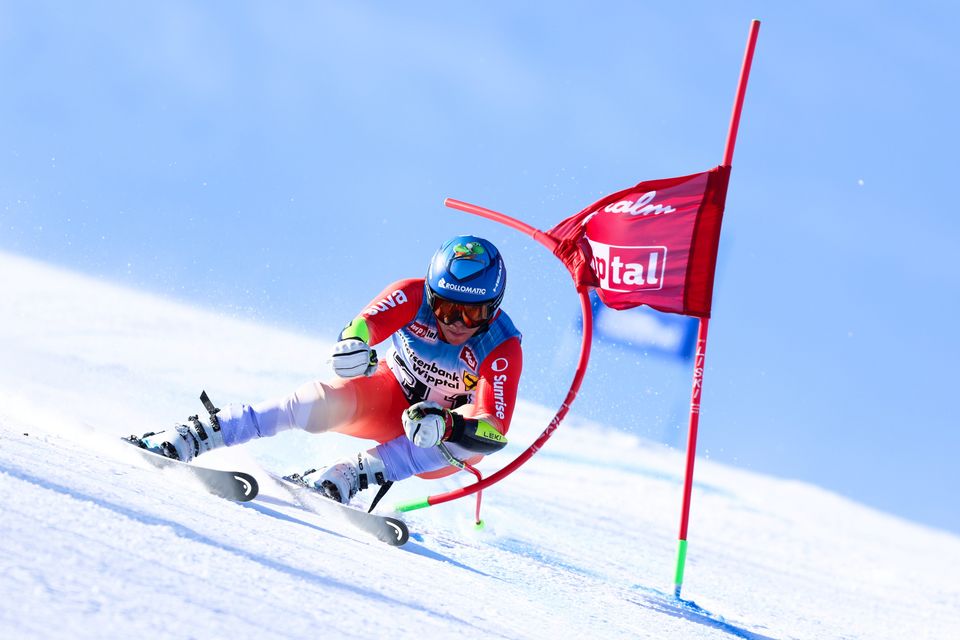 Ein Skifahrer in farbenfroher Rennkleidung und mit blauem Helm macht während eines Skirennens eine scharfe Kurve um ein rotes Slalomtor auf einer verschneiten Piste, wobei Schnee von seinen Skiern aufspritzt.