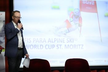 Ein Mann in einem blauen Blazer spricht in ein Mikrofon, während er Papiere in der Hand hält. Er steht vor einem projizierten Bild und dem Text "FIS Para Alpine Ski World Cup St. Moritz", während im Hintergrund ein Skifahrer fährt.