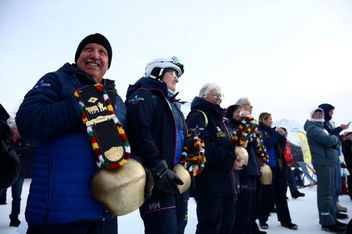 Eine Gruppe von Menschen in Winterkleidung steht in einer Reihe im Freien, jeder hält eine große Kuhglocke aus Messing, die mit bunten Bändern verziert ist. Der Boden ist mit Schnee bedeckt und im Hintergrund sind Berge zu sehen.