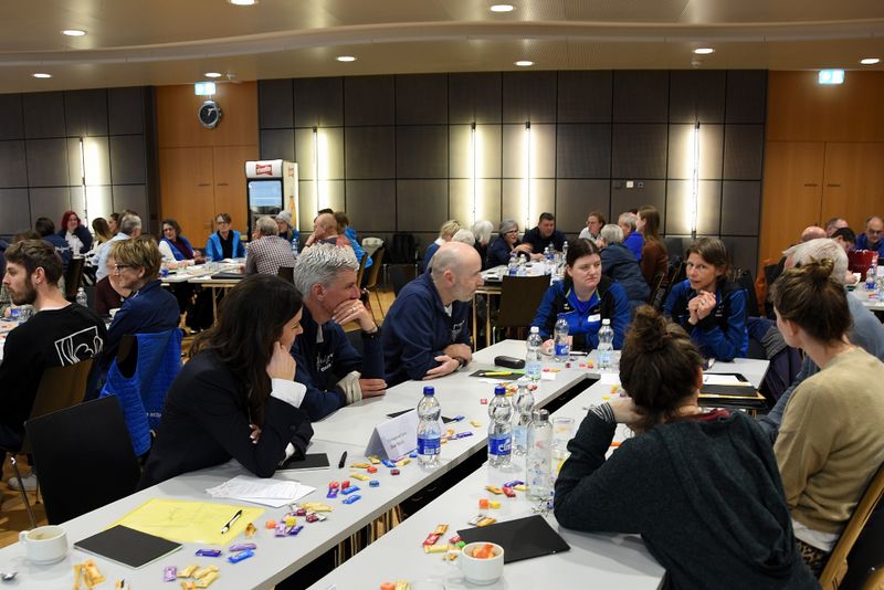 Eine Gruppe von Menschen sitzt in einem Konferenzraum an Tischen und diskutiert. Papiere, Getränke und bunte Süßigkeiten sind auf den Tischen verteilt. Im Hintergrund sind weitere Teilnehmer zu sehen, die eine geschäftige, gemeinschaftliche Atmosphäre schaffen.