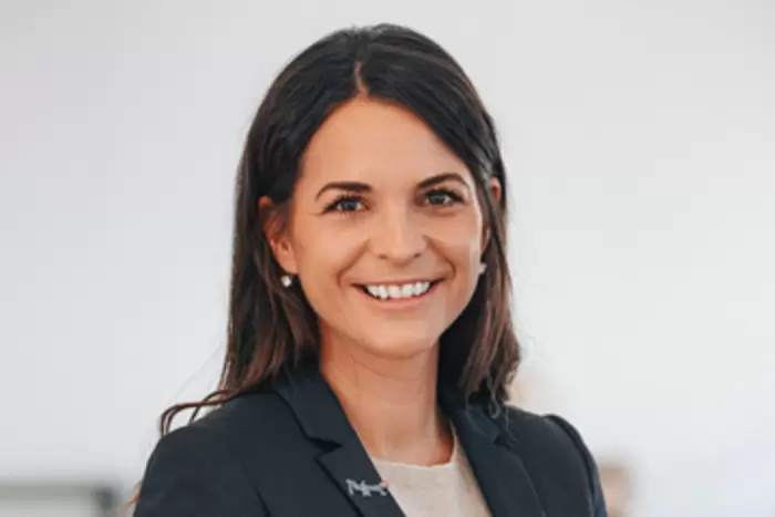 Eine Frau mit langen braunen Haaren, einem dunklen Blazer und einem hellen Oberteil trägt, lächelt und blickt vor einem schlichten, hellen Hintergrund in die Kamera.