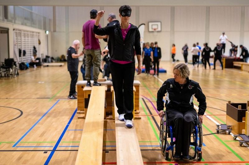 Eine Frau in einem Rollstuhl und mit einer Augenbinde steht auf einer Bank.