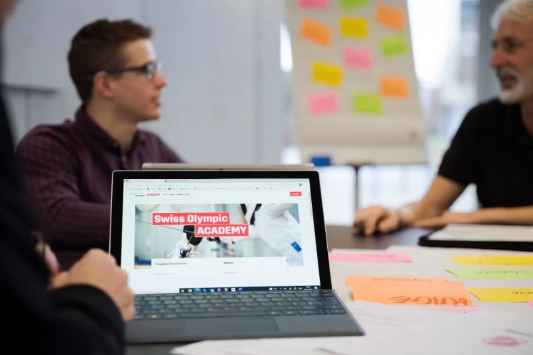Drei Personen sitzen an einem Tisch mit bunten Klebezetteln und führen eine Diskussion. Ein Laptop im Vordergrund zeigt die Website der Swiss Olympic Academy. Im Hintergrund befindet sich ein Flipchart mit Haftnotizen.
