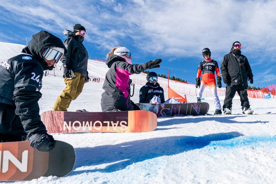 Eine Gruppe von Snowboardern sitzt und steht auf einem verschneiten Hang, ausgestattet mit Helmen und Winterbekleidung sowie bunten Snowboards. Eine Person zeigt den Hang hinauf. Im Hintergrund sind ein strahlend blauer Himmel und eine Rennstrecke zu sehen.