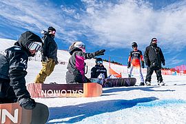 Eine Gruppe von Snowboardern sitzt und steht auf einem verschneiten Hang, ausgestattet mit Helmen und Winterbekleidung sowie bunten Snowboards. Eine Person zeigt den Hang hinauf. Im Hintergrund sind ein strahlend blauer Himmel und eine Rennstrecke zu sehen.
