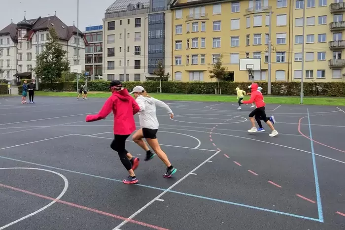 Die meisten Spieler tragen Jacken und Mützen, was auf kühles Wetter hindeutet. Im Hintergrund sind mehrere Gebäude mit vielen Fenstern zu sehen.