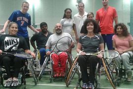 Eine bunt gemischte Gruppe von zehn Personen, darunter vier Rollstuhlfahrer, posiert gemeinsam auf einem Tennisplatz, hält Schläger in der Hand und lächelt in die Kamera.