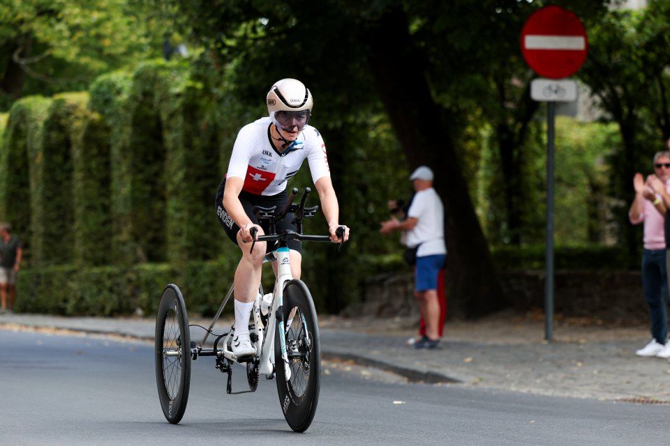 Ein Radfahrer, der einen weißen Helm und ein Schweizer Trikot trägt, fährt mit einem dreirädrigen Handbike auf einer Straße, mit Zuschauern und Grün im Hintergrund an einem sonnigen Tag.
