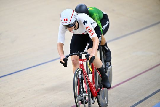 Zwei Radfahrer fahren ein Rennen auf einer Velodrombahn. Der vordere Fahrer trägt eine weiß-schwarze Uniform mit "SUI" auf der Brust und einen weißen Helm mit einem roten Kreuz. Der hintere Radfahrer trägt eine grün-schwarze Uniform und einen Helm.