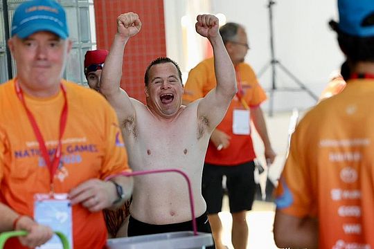 Ein fröhlicher, hemdsärmeliger Mann mit erhobenen Armen steht inmitten anderer Menschen, die orangefarbene Hemden tragen, bei einer scheinbaren Sportveranstaltung.