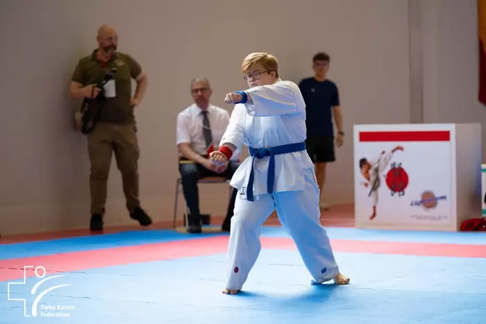 Ein junger Karate-Athlet in weißem Gi und blauem Gürtel führt eine Kata auf einer Matte aus. Im Hintergrund sind Offizielle und Beobachter zu sehen. In der unteren linken Ecke ist das Logo des Schweizerischen Karateverbands zu sehen.