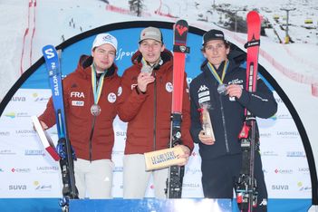 Drei männliche Skifahrer in Skikleidung stehen auf einem Podium, jeder hält eine Medaille und Skier in der Hand, mit einer verschneiten Piste und Veranstaltungsbannern im Hintergrund. Sie lächeln und freuen sich über ihren Sieg.