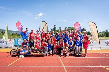 Eine bunte Gruppe von Sportlern, darunter auch Rollstuhlfahrer, posiert und feiert gemeinsam auf einer Laufbahn. Sie tragen rote und blaue Trikots, im Hintergrund sind Flaggen und ein strahlend blauer Himmel zu sehen.