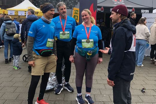 Drei Marathonläufer in blauen Hemden und mit Medaillen posieren für ein Foto und unterhalten sich mit einem Mann in Jacke und Mütze bei einer Veranstaltung im Freien mit Zelten und Menschen im Hintergrund.