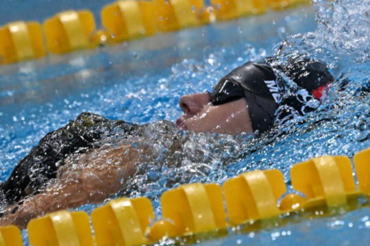 Ein Schwimmer mit Schwimmbrille und Badekappe nimmt an einem Rückenschwimmwettbewerb teil; er befindet sich teilweise unter Wasser in einem Becken mit gelben Bahngrenzen, und ihm spritzt Wasser ins Gesicht.