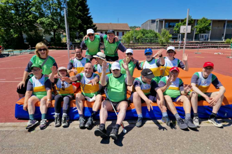 Eine Gruppe von Menschen in Sportkleidung und grünen Westen sitzt im Freien auf Matten und lächelt und winkt der Kamera auf einem Sportplatz bei sonnigem Wetter zu.