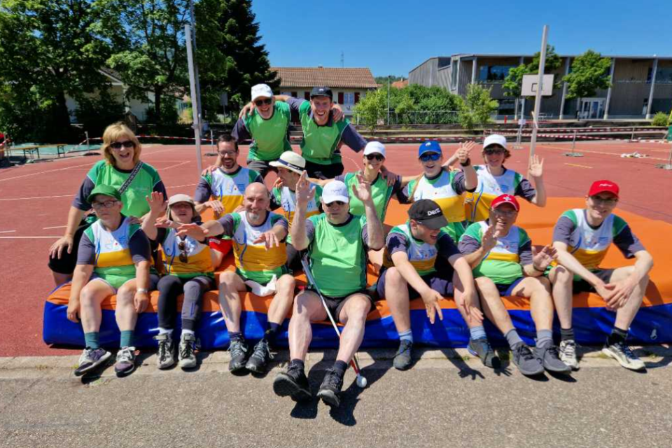 Eine Gruppe von Menschen in Sportkleidung und grünen Westen sitzt im Freien auf Matten und lächelt und winkt der Kamera auf einem Sportplatz bei sonnigem Wetter zu.