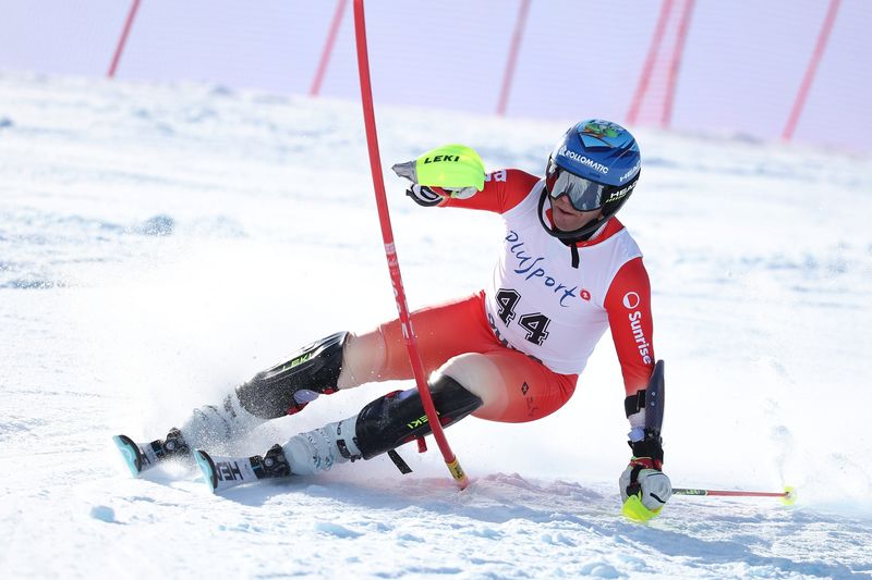 Ein Skifahrer in rot-weißer Rennkleidung, mit blauem Helm und gelben Handschuhen, macht eine scharfe Kurve um ein Slalomtor auf einer verschneiten Strecke und wirft dabei Schnee auf, während er sich dicht an den Boden lehnt.