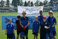 Eine Gruppe junger Sportler in blauen Trikots posiert vor einem Fußballtor mit einem Plusport-Banner. Einige tragen Mützen, und alle halten blaue Kordeltaschen in der Hand. Sie befinden sich an einem sonnigen Tag im Freien.