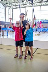 Drei lächelnde Kinder in Sportkleidung feiern auf einem Hallenplatz und halten mit erhobenen Armen Floorballschläger in den Händen. Im Hintergrund sind bunte Banner und Sportgeräte zu sehen.