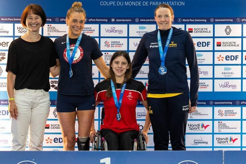 Vier Frauen, von denen drei Medaillen tragen, posieren und lächeln auf einem Podium bei einer Para-Schwimmveranstaltung. Eine Frau in der Mitte benutzt einen Rollstuhl. Auf dem Hintergrund hinter ihnen sind Sponsorenlogos zu sehen.