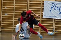 Zwei Hallenfußballspieler stoßen zusammen; einer in rot-schwarzer Kleidung fällt über einen Spieler in Weiß, der auf dem Boden liegt. Ein Fußball liegt in ihrer Nähe. Im Hintergrund sind hölzerne Sprossenwände und ein Banner zu sehen.