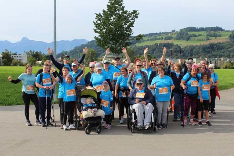 Eine große Gruppe von Menschen in blauen Hemden, einige mit nummerierten Lätzchen, lächeln und winken im Freien. Einige benutzen Gehstöcke, eine Person sitzt im Rollstuhl und ein Kind ist in einem Kinderwagen unterwegs. Im Hintergrund sind grüne Hügel und Bäume zu sehen.