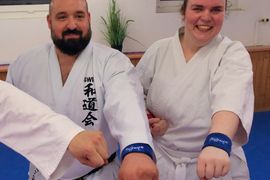 Zwei Personen in weißen Karateuniformen lächeln in die Kamera. Sie stehen nebeneinander und zeigen blaue Armbänder an ihren Fäusten, während der Arm einer weiteren Person im Vordergrund zu sehen ist. Im Hintergrund ist eine Pflanze zu sehen.