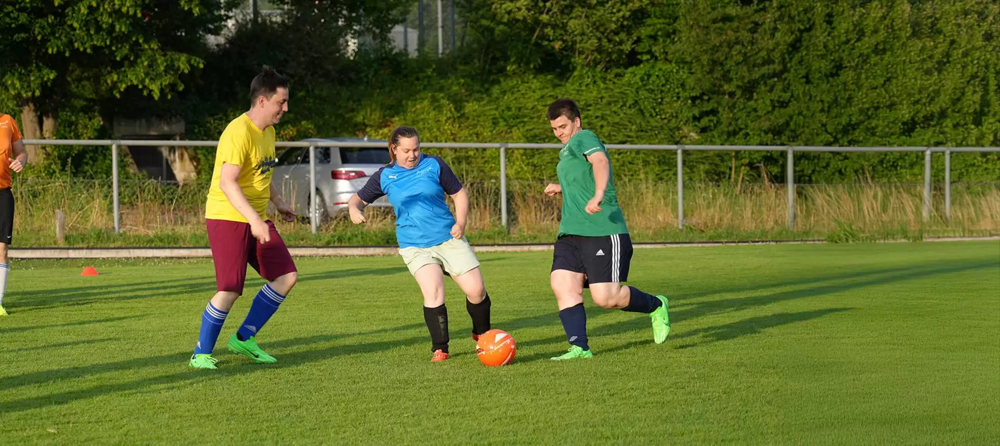Drei Personen spielen Fußball auf einer Wiese; eine in Gelb, eine in Blau und eine in Grün kämpfen um einen orangefarbenen Ball. Im Hintergrund sind Bäume und ein Zaun zu sehen.