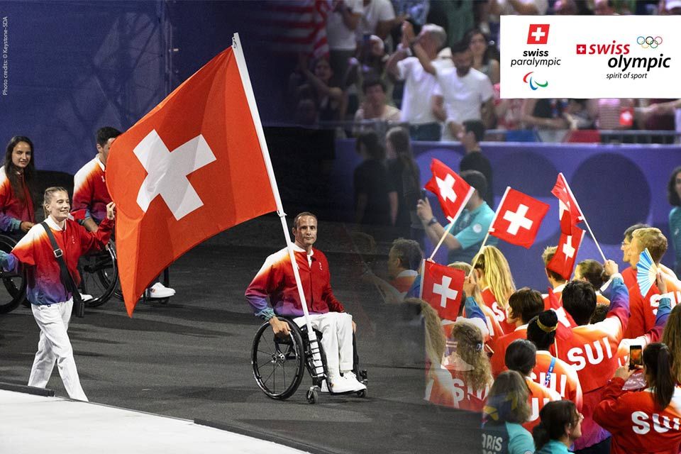 Ein lächelnder Schweizer Paralympics-Athlet im Rollstuhl trägt stolz eine große Schweizer Flagge bei einem Hallen-Event, umgeben von Teamkollegen in roten Uniformen. In der Ecke sind die Logos der Schweizer Paralympics und der Olympischen Spiele zu sehen.