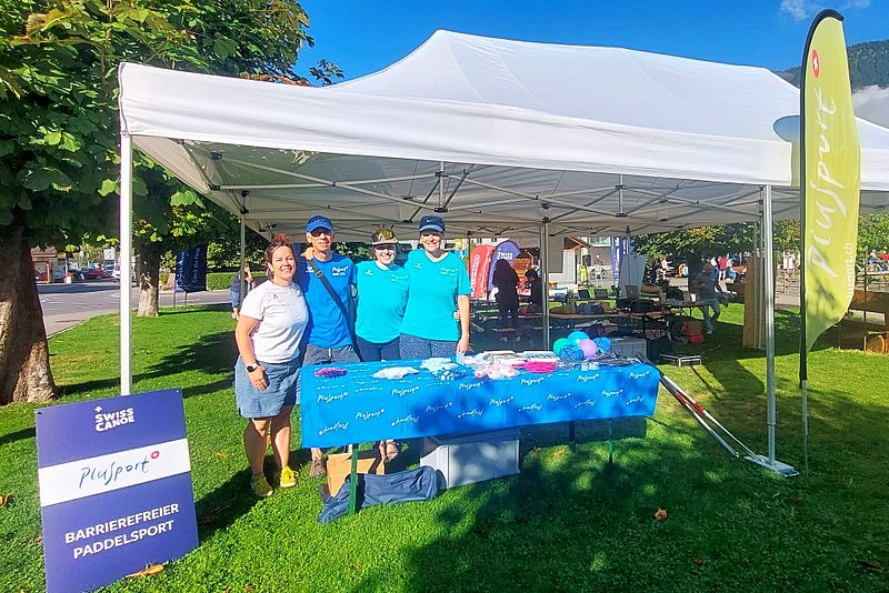 Vier Menschen stehen lächelnd vor einem Tisch mit blauem Tischtuch und Werbeartikeln unter einem weißen Zelt. Daneben steht auf einem Schild „Barrierefreier Paddelsport“. Im Hintergrund sind Bäume und ein klarer Himmel zu sehen.