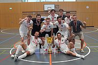Eine jubelnde Hallenfußballmannschaft in weißen Trikots posiert mit einem großen goldenen Pokal und Medaillen auf einem Sporthallenfeld und feiert ihren Sieg.