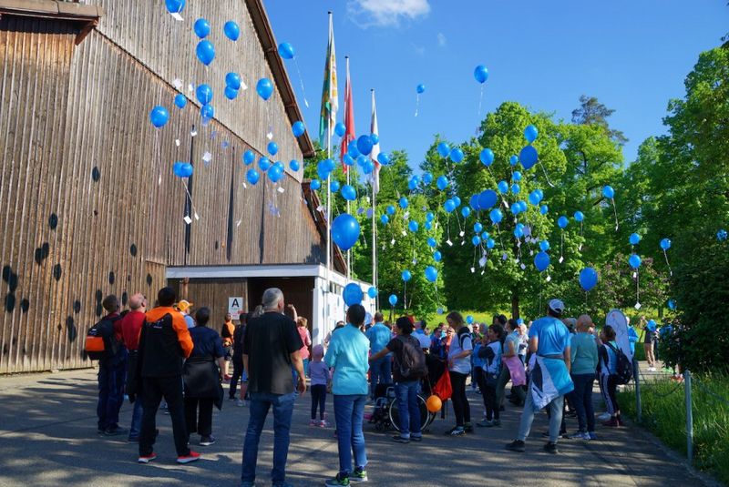 Eine Gruppe von Menschen hat sich im Freien versammelt und lässt neben einem Holzgebäude blaue Ballons in den Himmel steigen. Im Hintergrund sind an einem sonnigen Tag mehrere Flaggen und Bäume zu sehen.