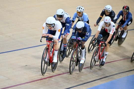 Eine Gruppe von Radfahrern in Rennkleidung und mit Helm tritt in einem Indoor-Velodrom gegeneinander an und fährt eng zusammen auf der Holzbahn.