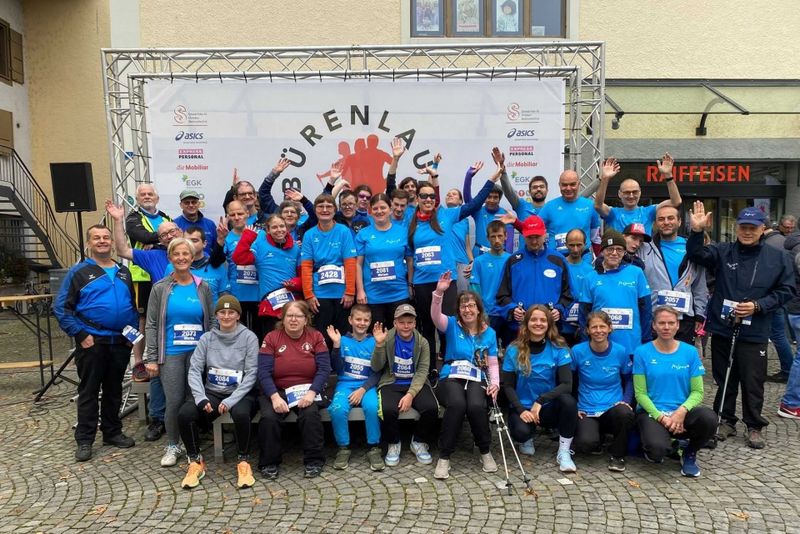 Eine große Gruppe von Menschen in blauen Hemden und Sportkleidung posiert und lächelt gemeinsam vor einer Kulisse für ein Rennen, einige halten Startnummern in der Hand. Der Schauplatz scheint im Freien auf einer Kopfsteinpflasterfläche zu sein.