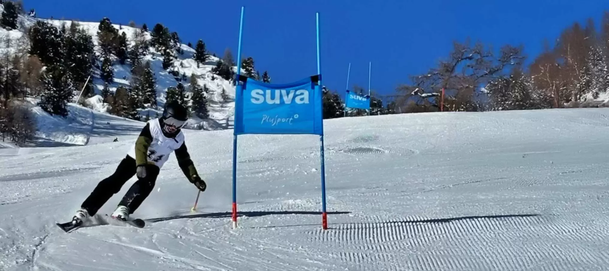 Ein Skifahrer, der einen Helm und eine Skibrille trägt, fährt auf einer verschneiten Piste bergab und manövriert um ein blaues Tor mit der Aufschrift suva unter einem klaren blauen Himmel mit Bäumen und Bergen im Hintergrund.